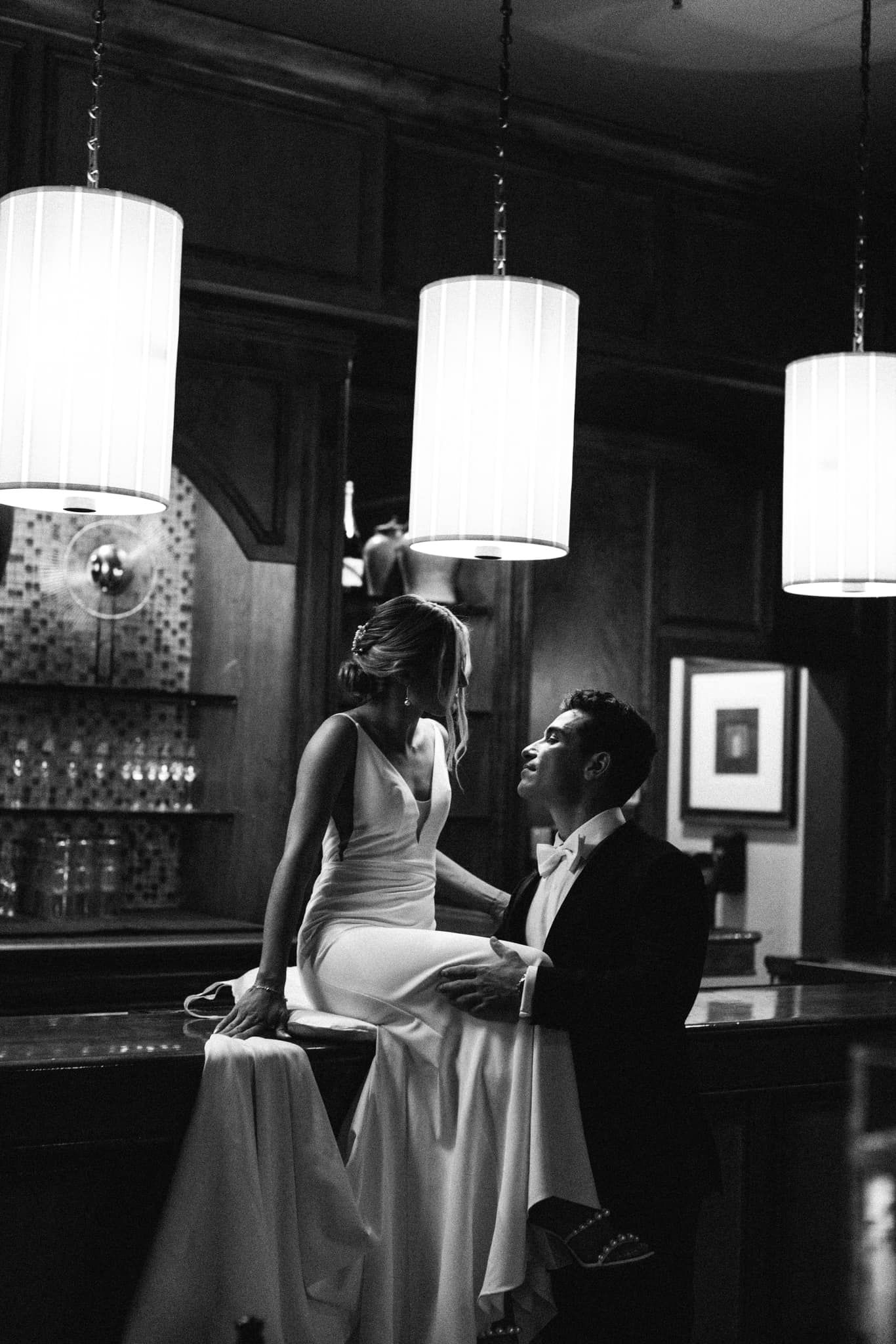 Couple in formal attire share romantic moment at bar with pendant lights.
