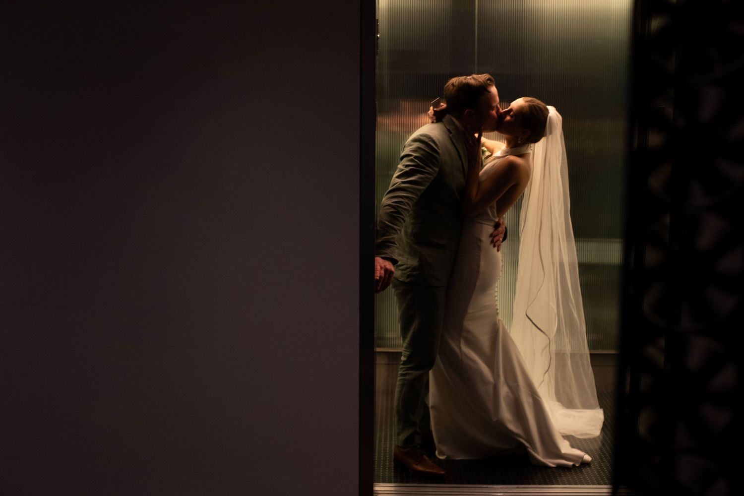 A romantic wedding moment captured in an elevator with a bride in white gown and veil kissing her groom.