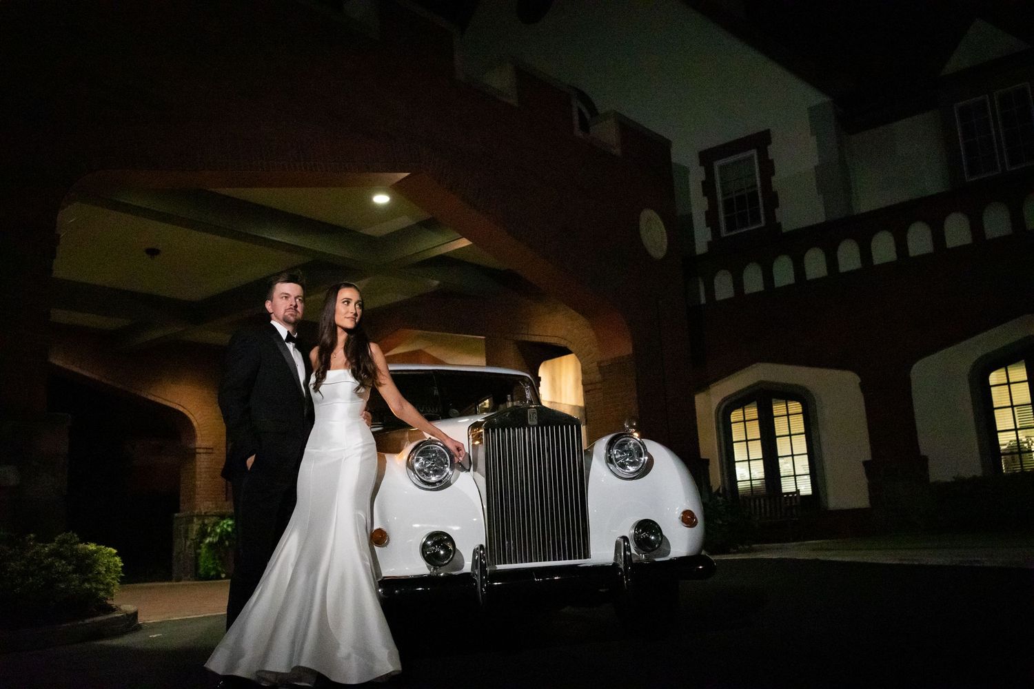 Elegant couple poses with vintage Rolls Royce at night outside historic venue.