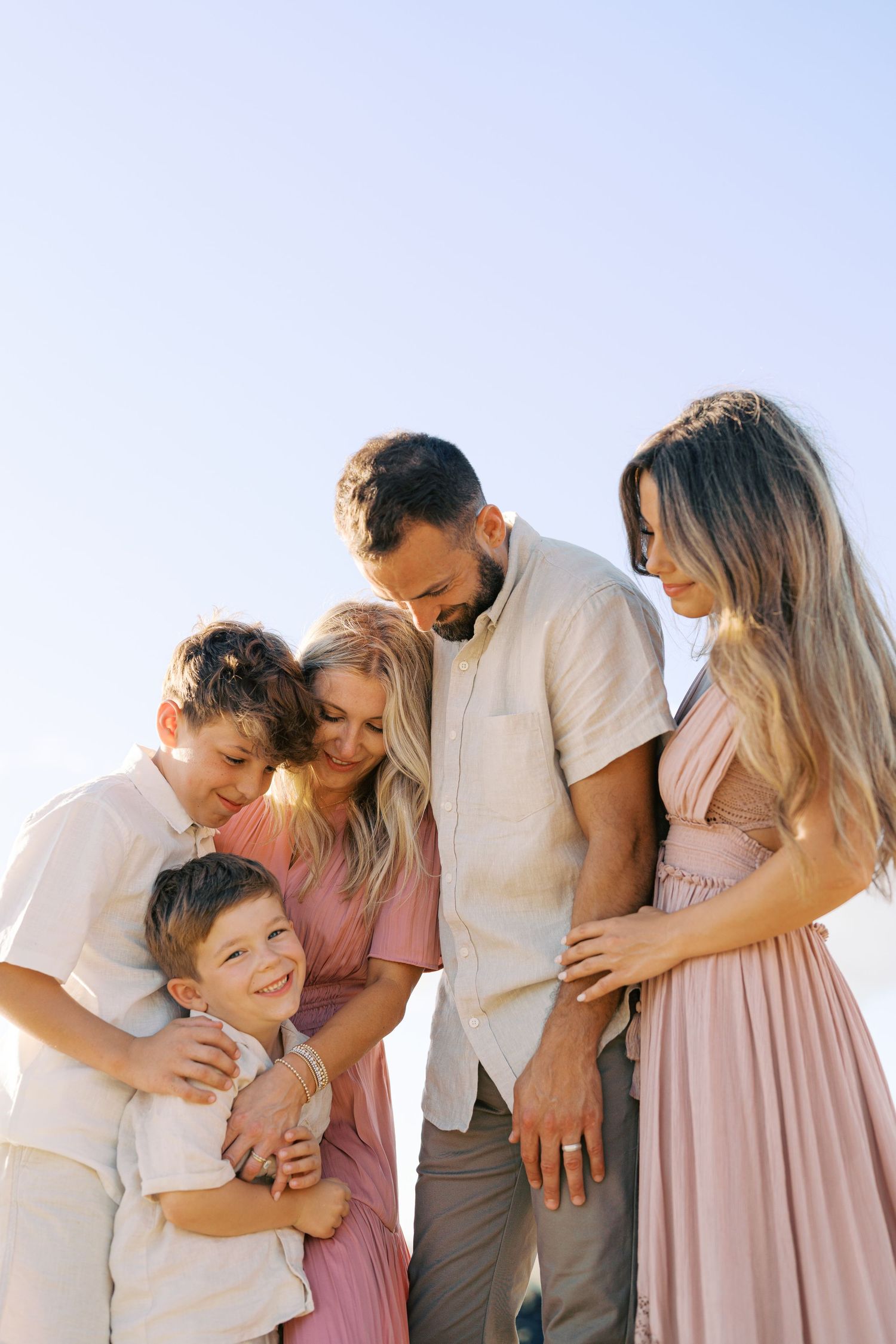 A family of five embraces while wearing coordinated neutral outfits against blue sky background.