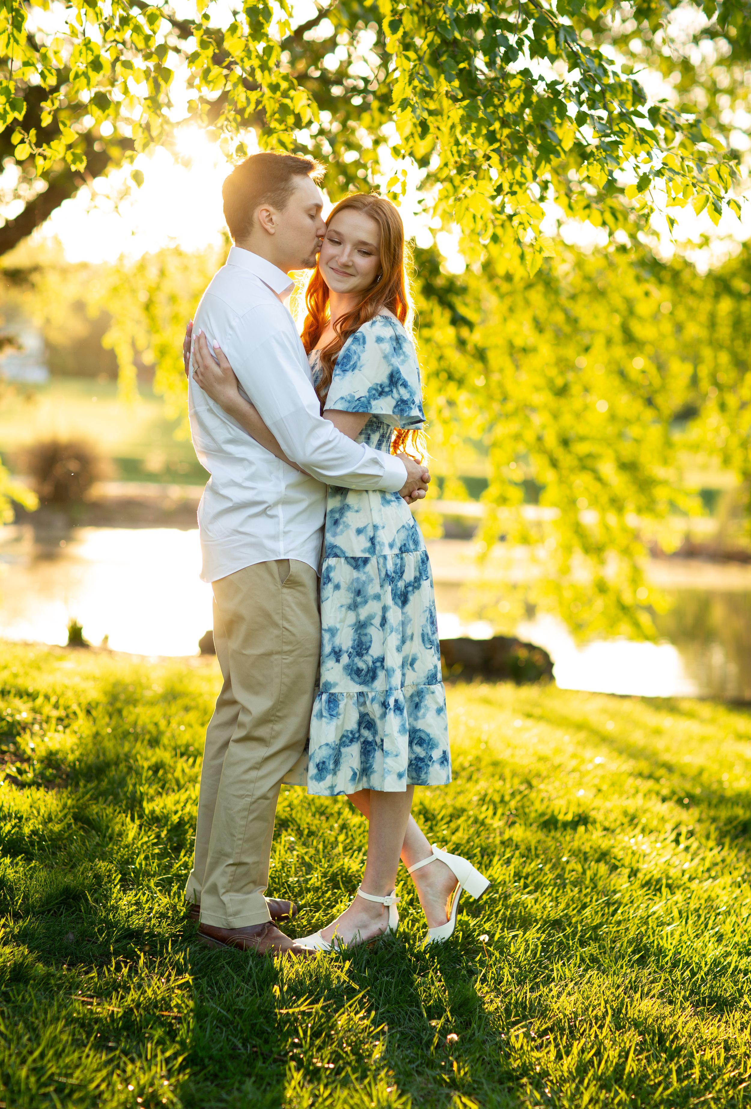 Couple shares a tender embrace under weeping willow tree in golden hour engagement photo.