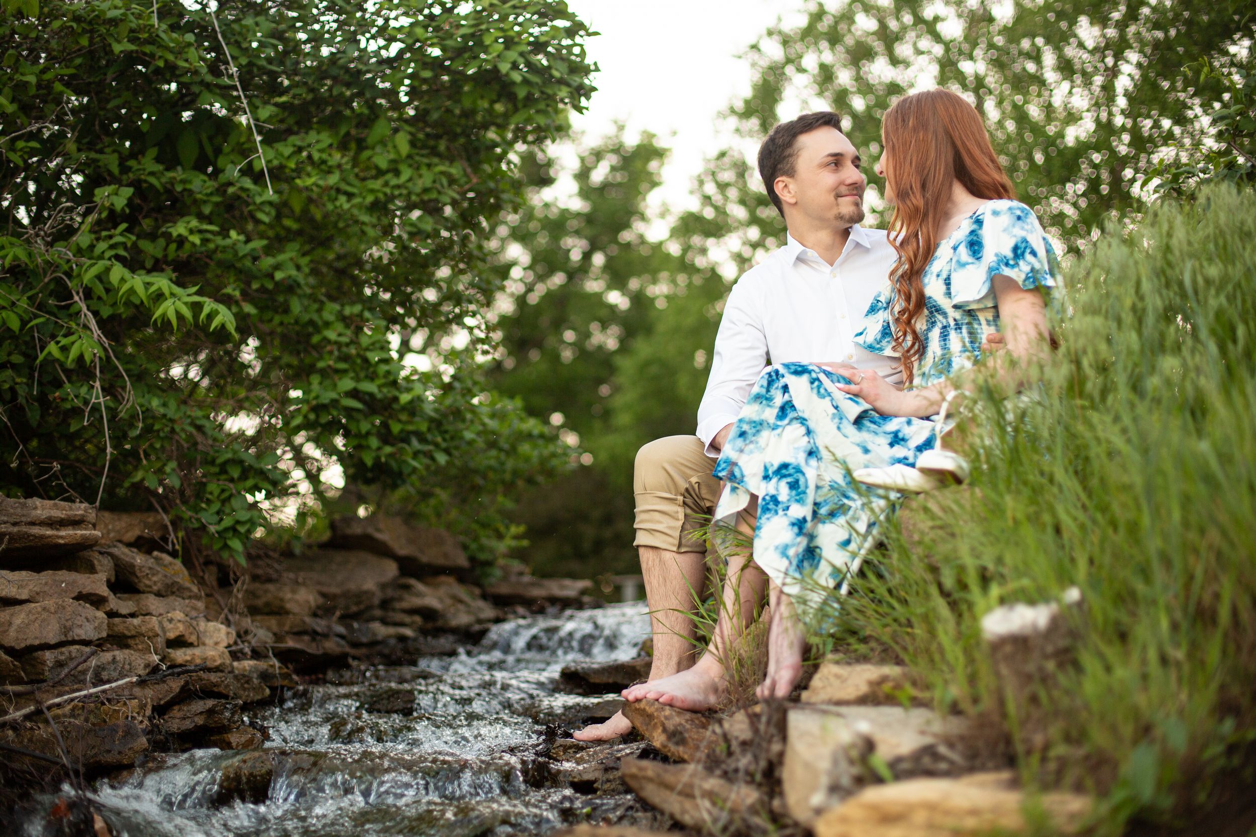 Engaged couple sits together on rocks beside a peaceful stream surrounded by lush greenery.