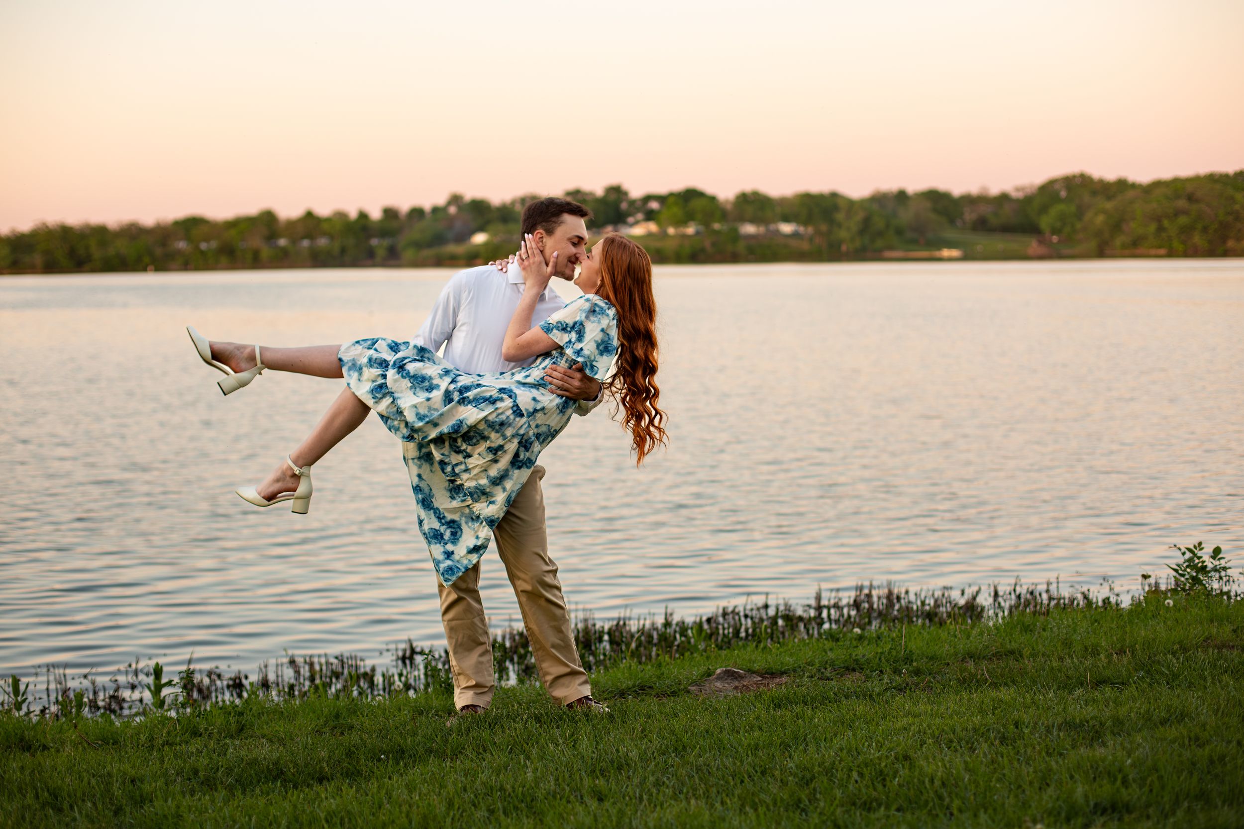 Man lifts and dips woman in floral dress during romantic lakeside sunset engagement photo.