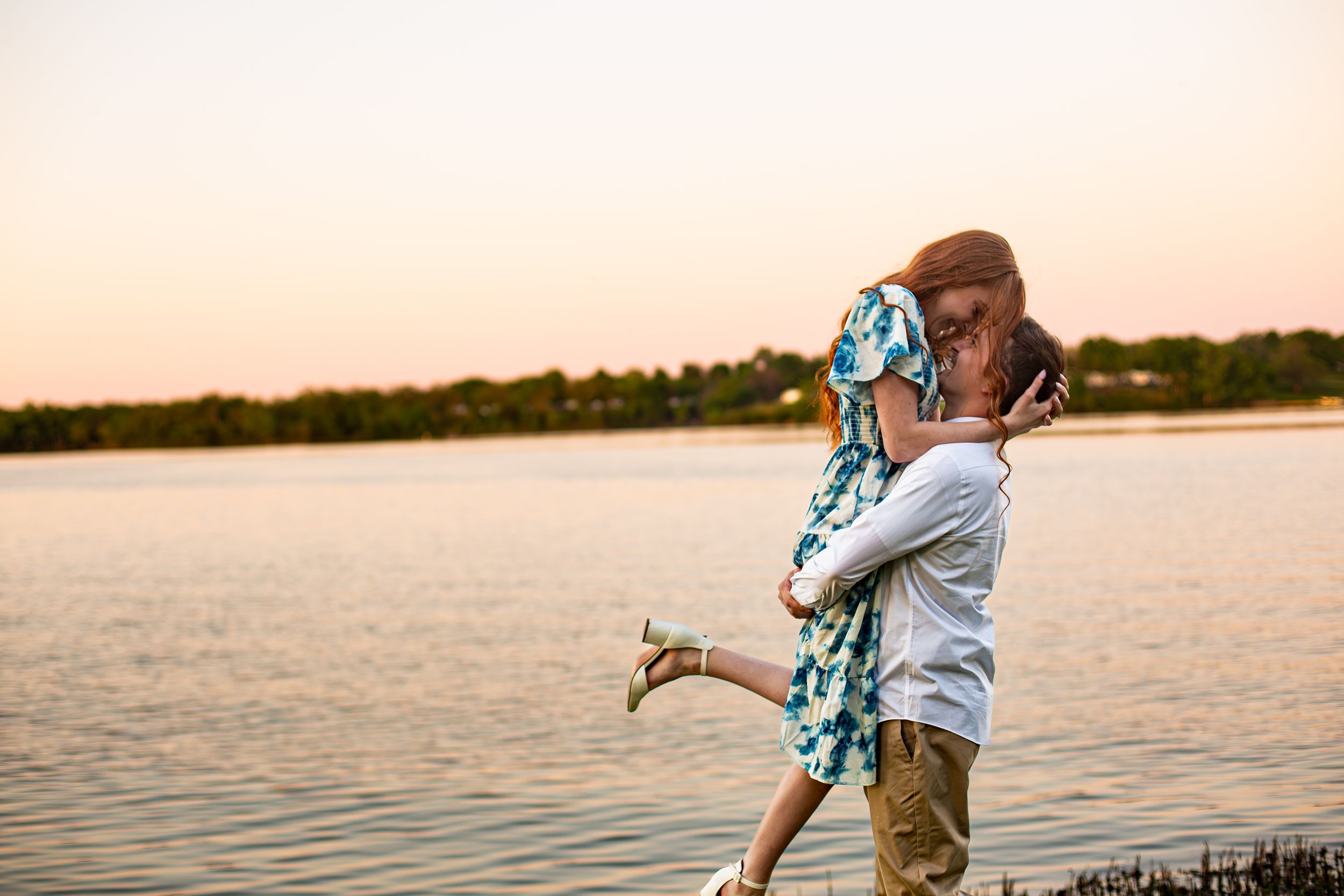 Couple shares a joyful kiss by the lake at sunset during outdoor engagement session.
