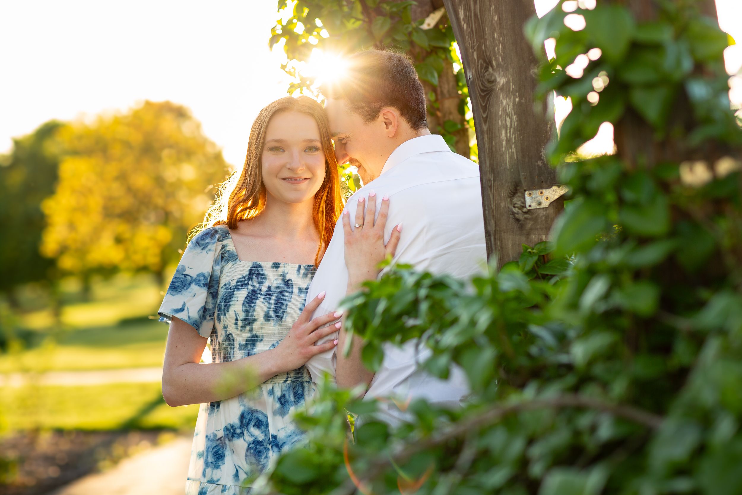 Couple embraces near ivy-covered tree in golden sunset light during outdoor engagement session.