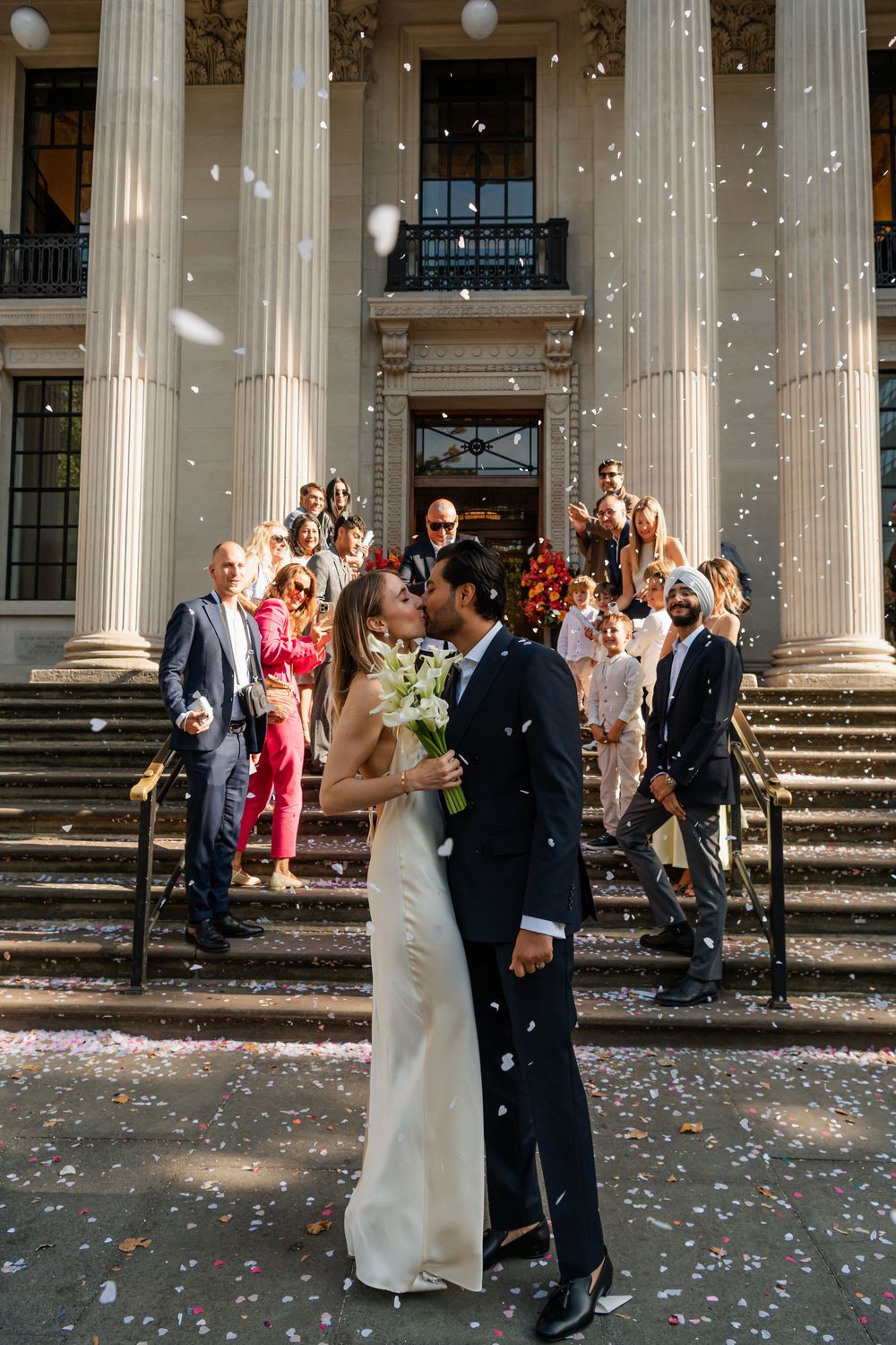 Wedding party gathered on grand steps of historic building with confetti floating in sunlight.