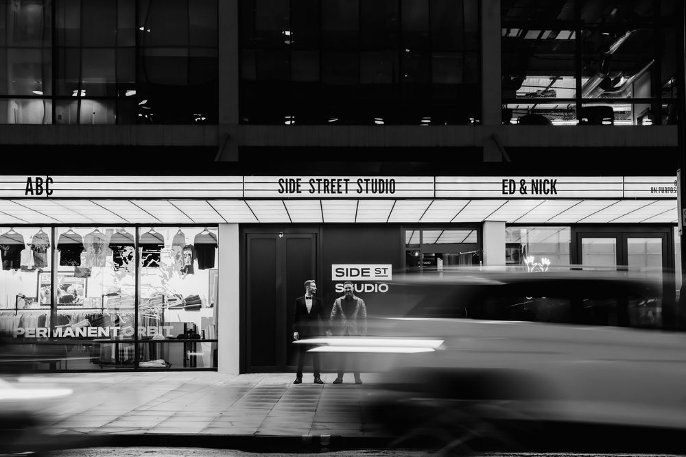 Black and white photo of a storefront at night with motion blur from passing car lights.