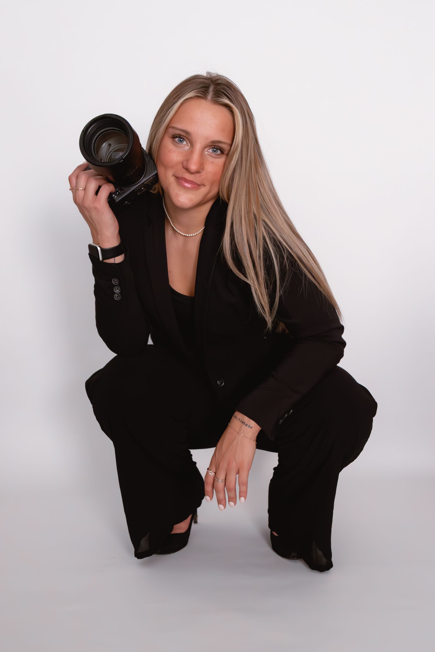 Professional photographer strikes dynamic poses in a black suit against a white background while holding a DSLR camera.