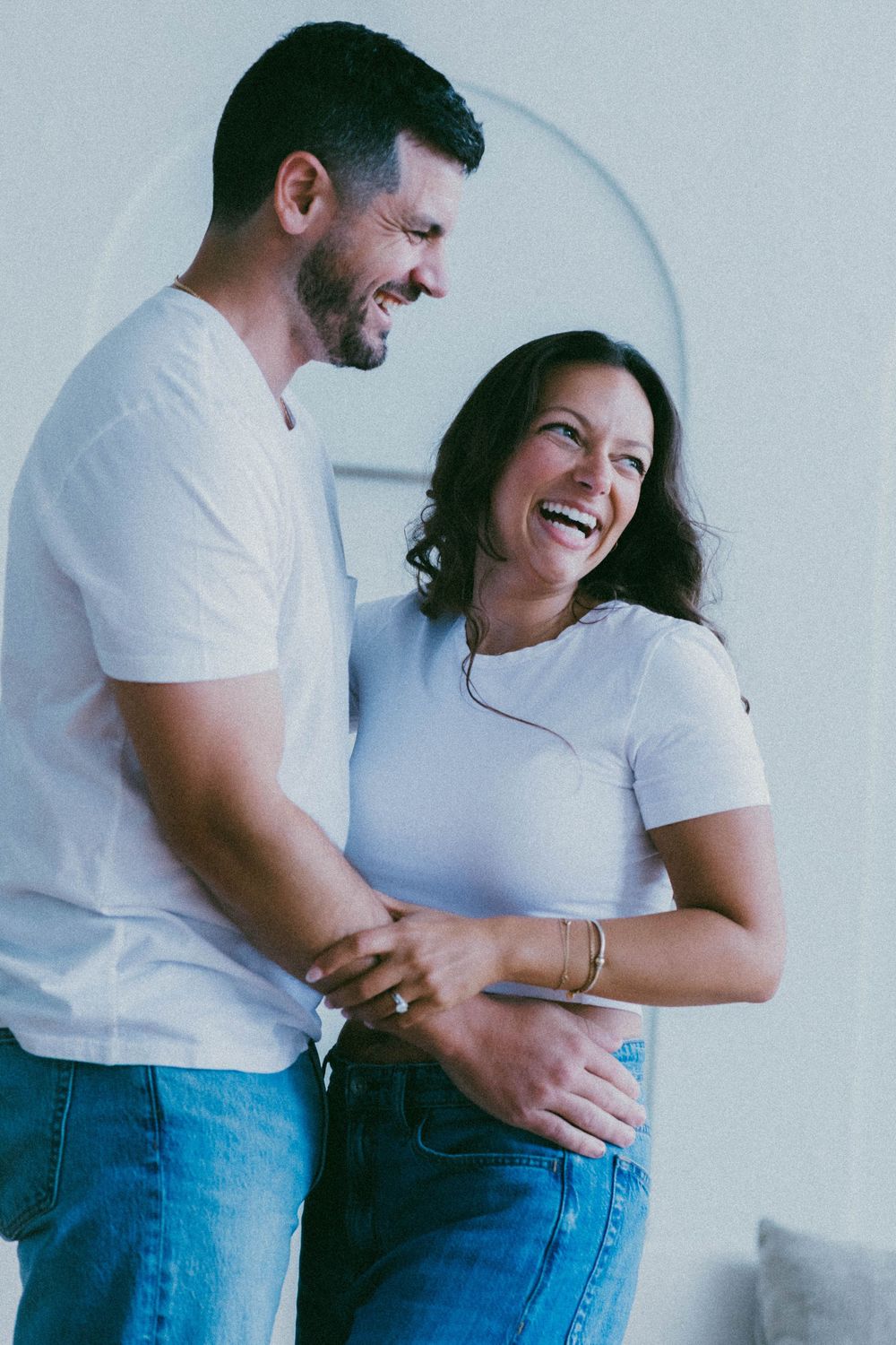 Laughing couple in matching white t-shirts and jeans embracing in a bright modern living room.