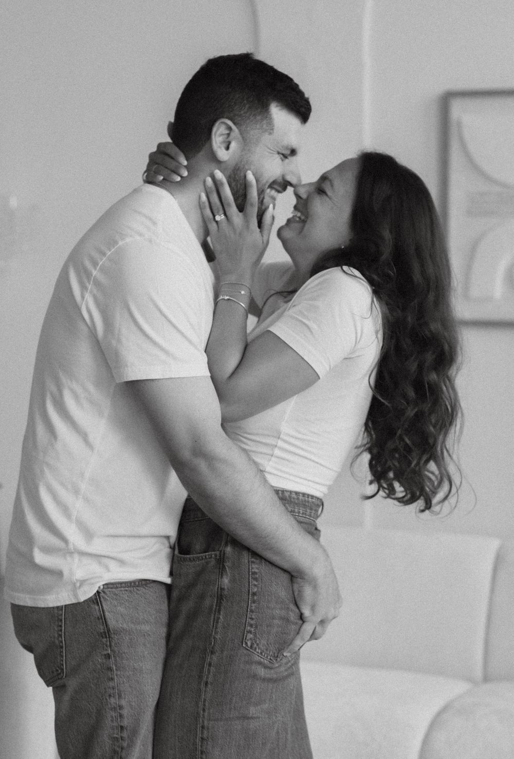 Black and white photo of a couple smiling and nearly kissing while embracing in a bright room.