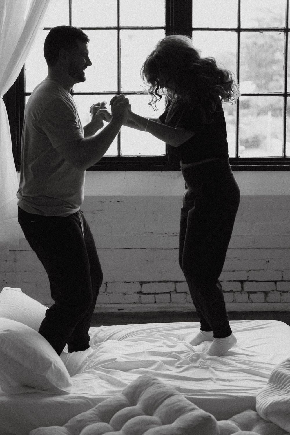 Black and white photo of a couple dancing and holding hands while jumping on a bed near large windows.
