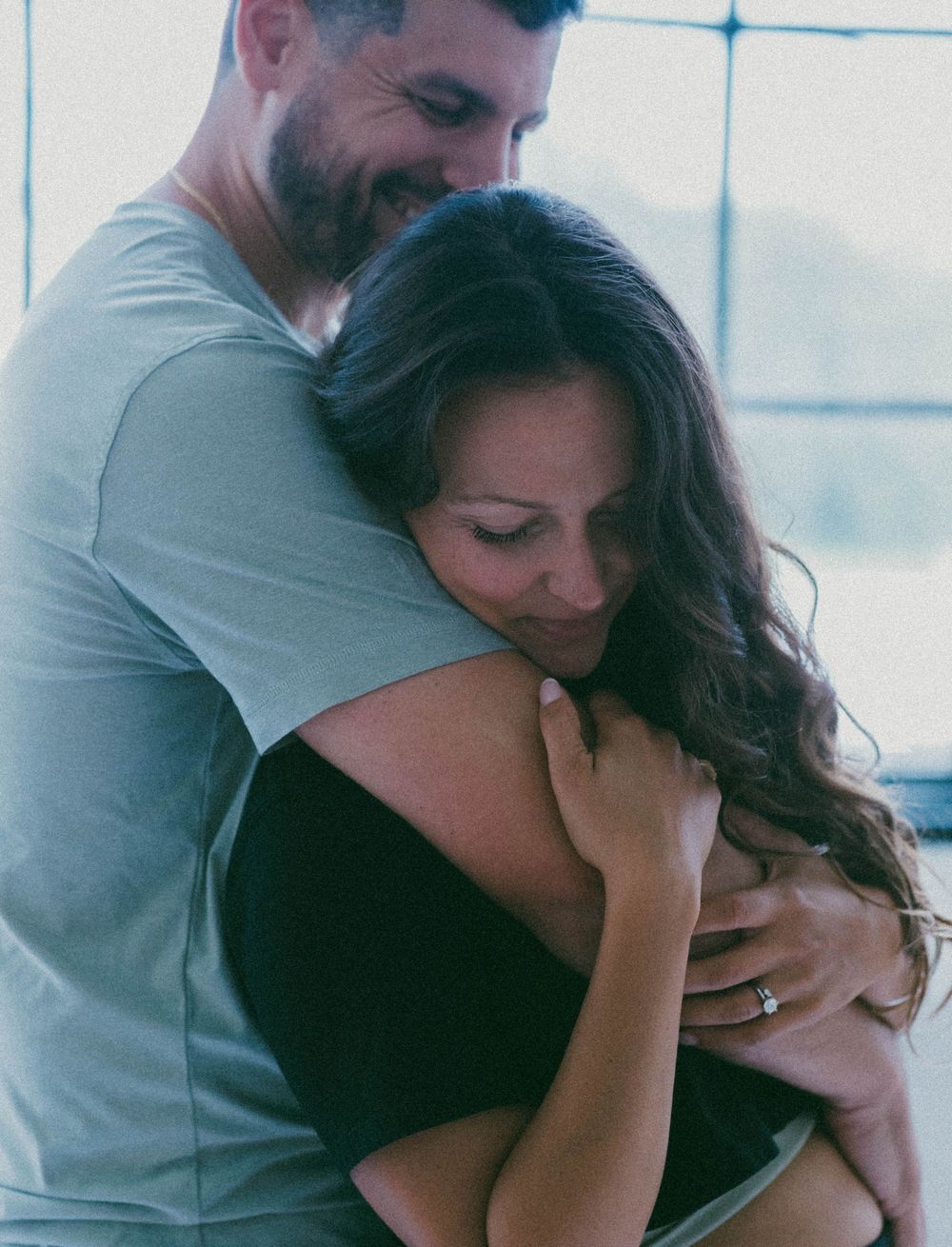 A smiling couple sharing a warm embrace, the woman wearing an engagement ring, in a softly lit room.