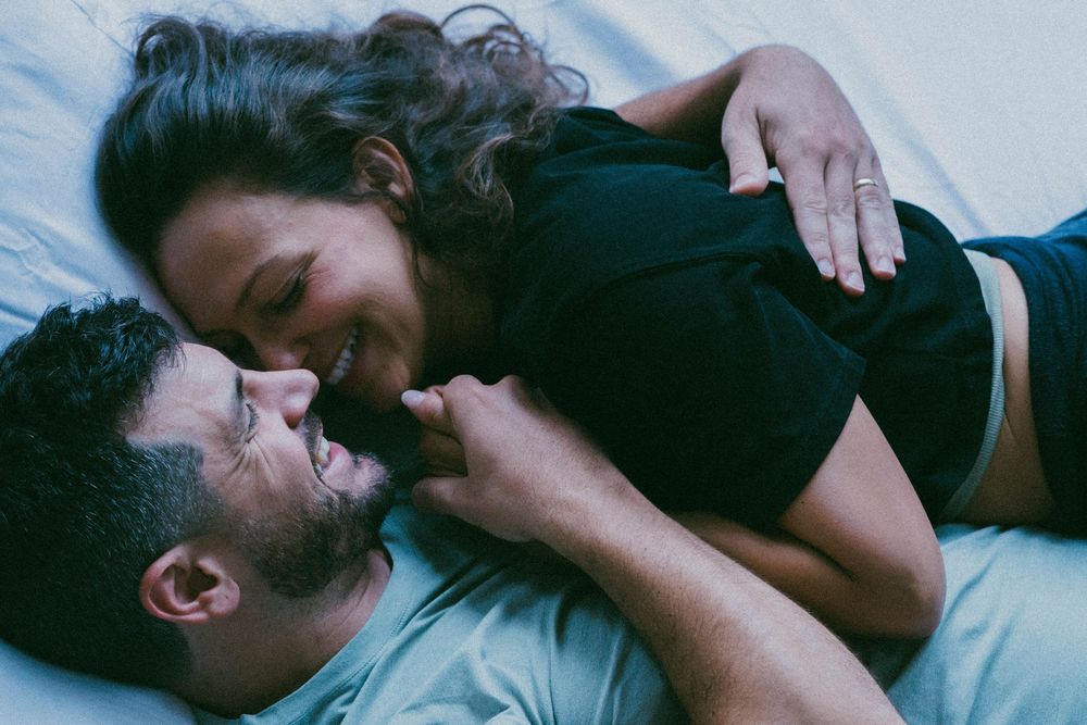 A couple lying on a bed, smiling and gazing at each other in a warm and intimate moment.