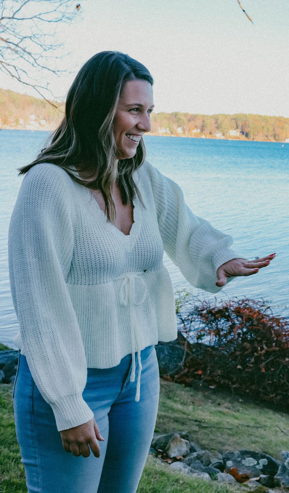 Smiling woman in white knit sweater and jeans standing by a lakeside on a cool autumn day.