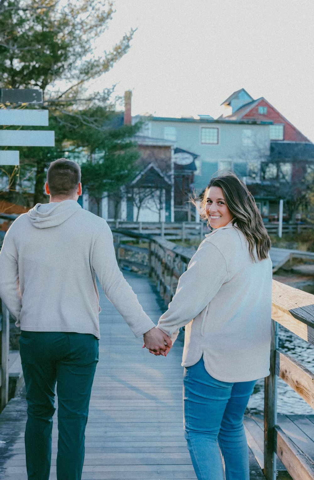 Happy couple holding hands walking on a wooden boardwalk near waterfront shops, woman looking back smiling.