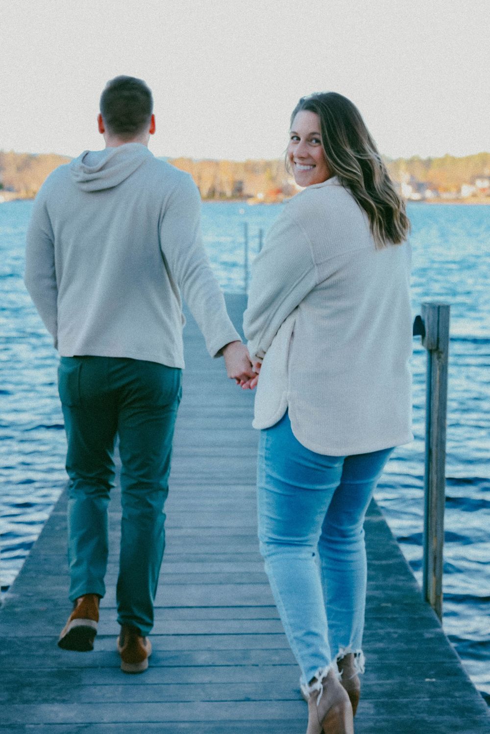 Couple holding hands on a dock by the water, woman in white jacket smiling back at camera.
