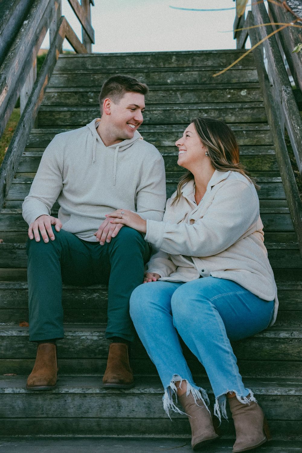 Smiling couple sitting on rustic wooden steps outdoors, wearing matching cream sweaters and jeans.