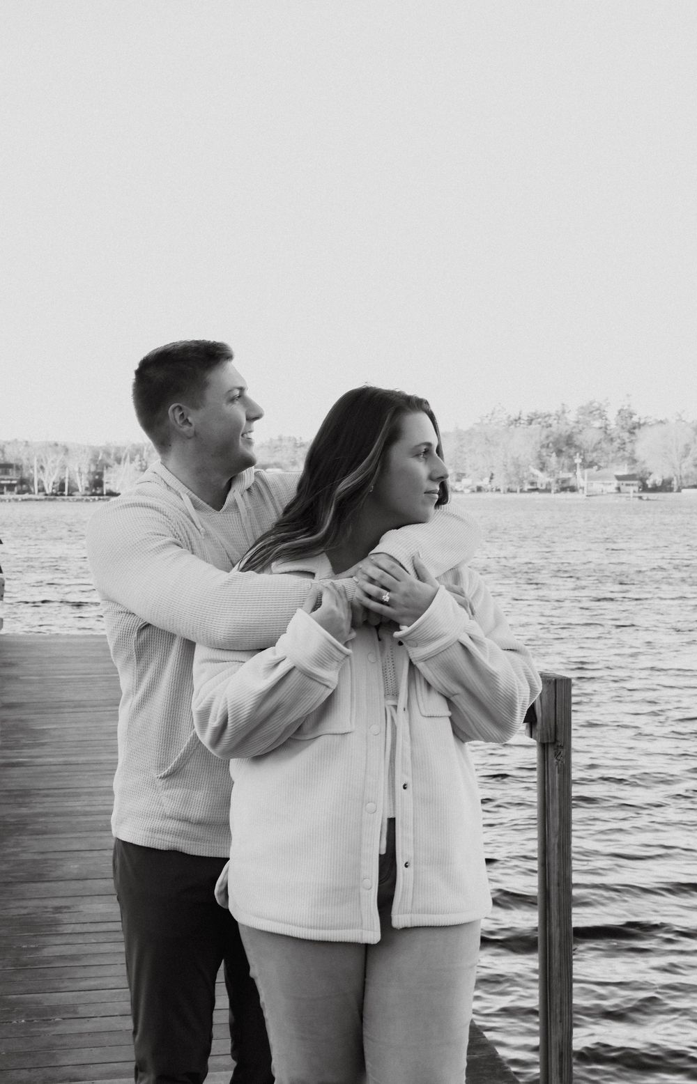 Black and white photo of man embracing woman from behind on a dock overlooking a calm lake.