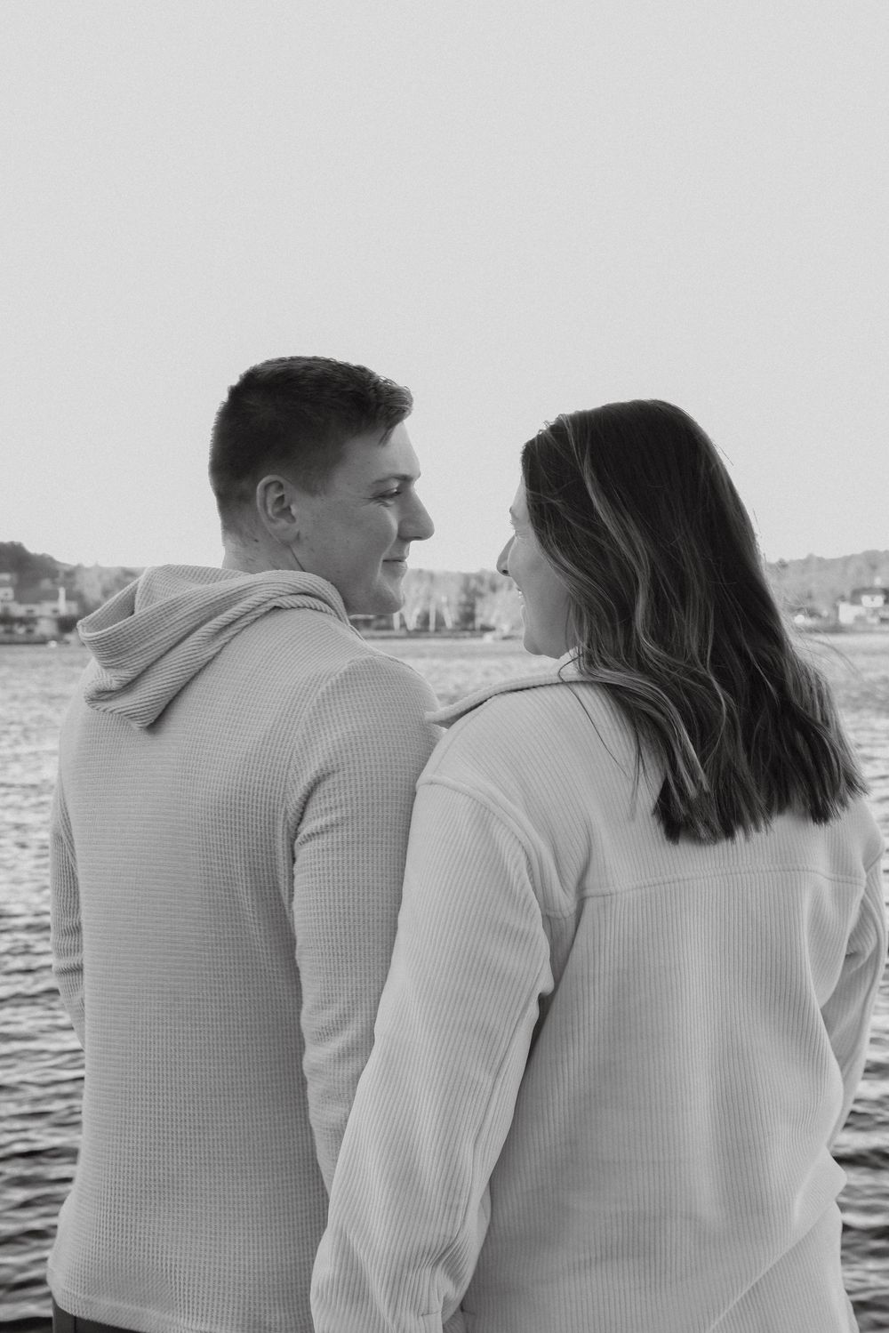 Black and white portrait of couple standing back to back by the water, smiling at each other.
