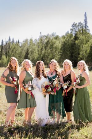 Wedding party poses in field with bridesmaids wearing olive green dresses holding colorful bouquets.