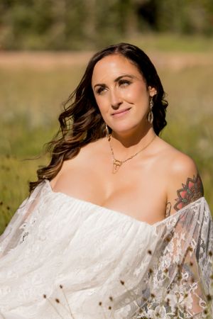 Bride in off-shoulder lace dress with visible tattoo poses outdoors in natural lighting.