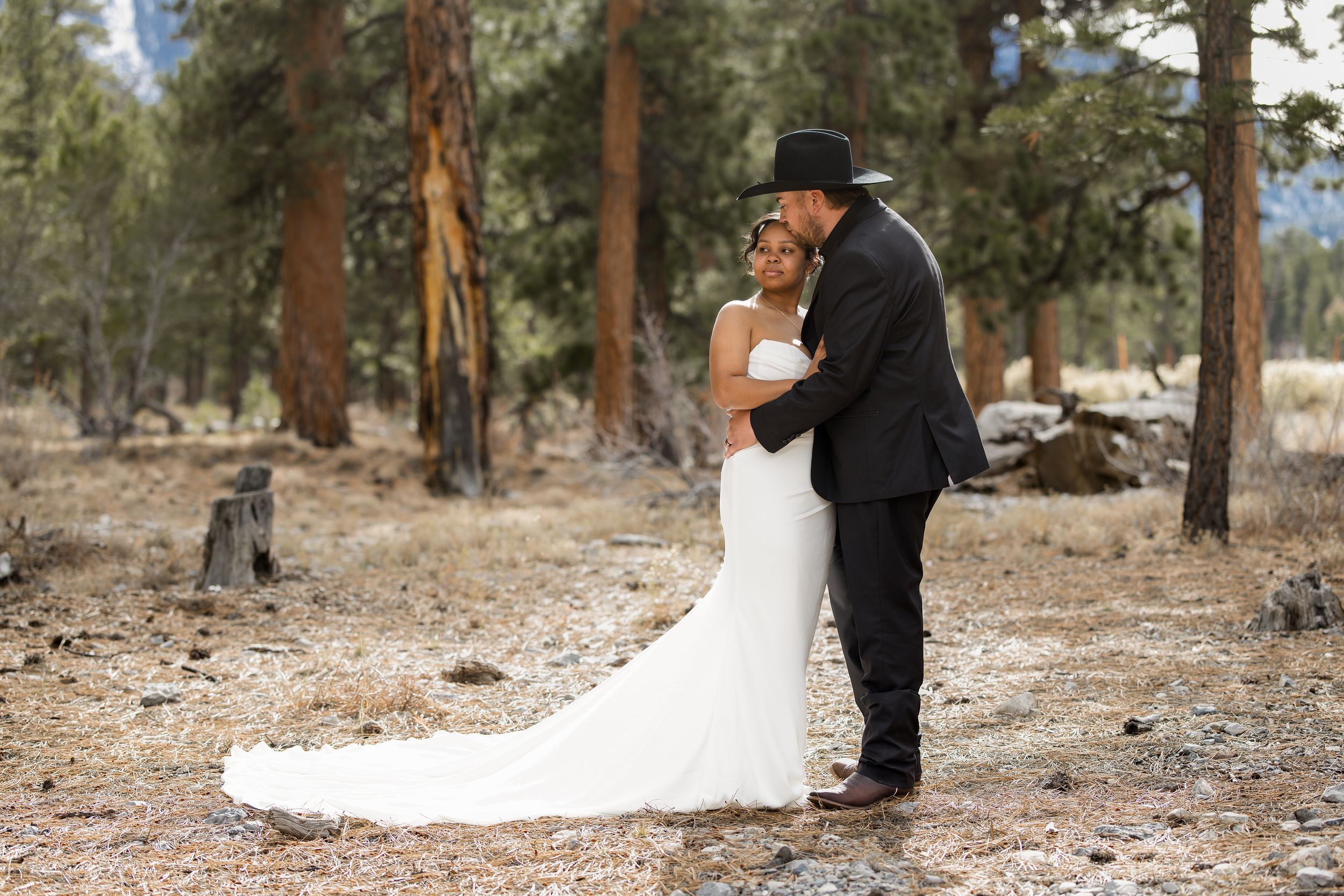 A couple in wedding attire embrace in a rustic forest setting with tall pine trees in the background.