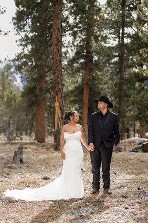 A couple in wedding attire walks together through a snowy pine forest during their winter wedding photoshoot.