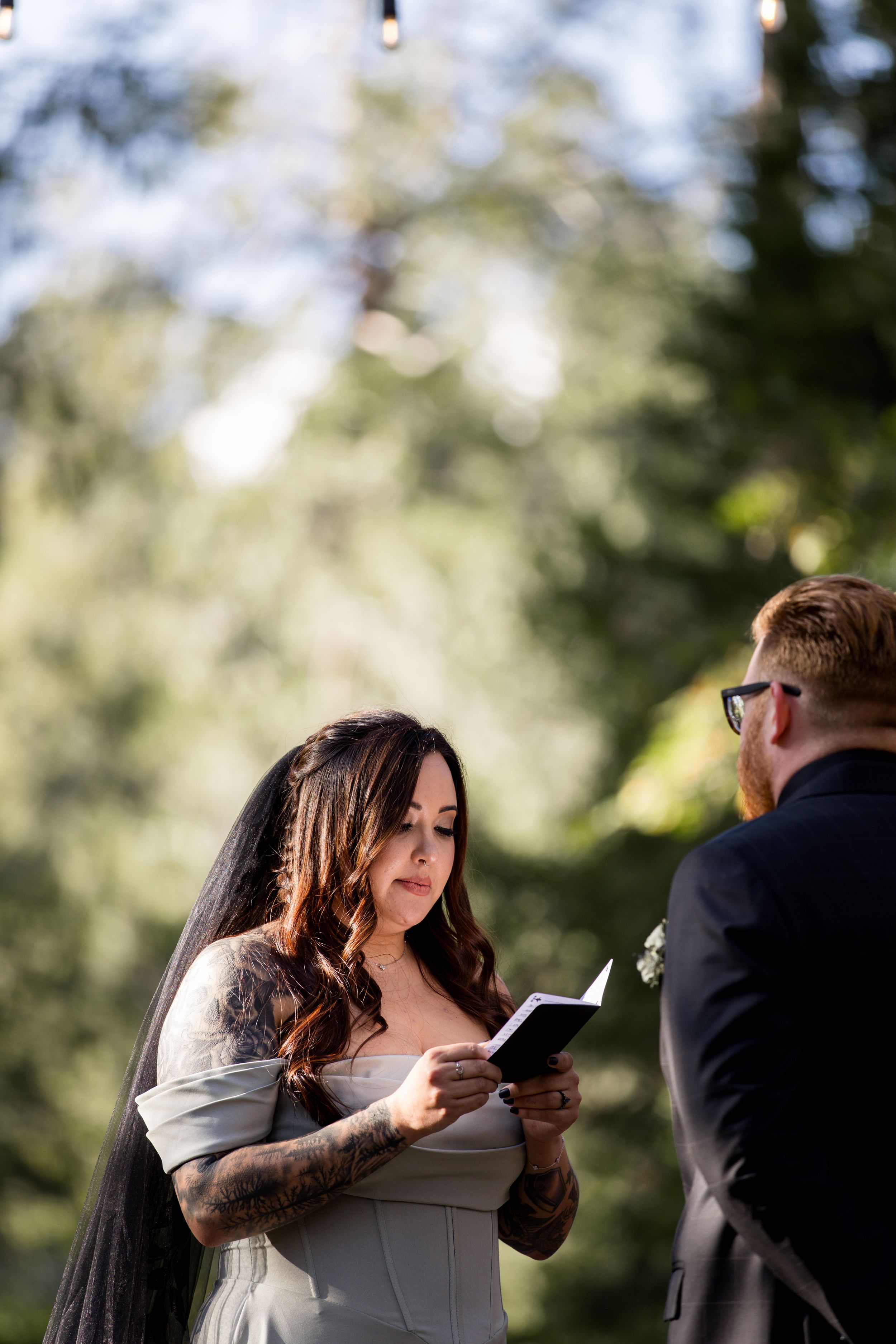 A couple exchanges vows during an outdoor wedding ceremony in a sunlit forest setting.