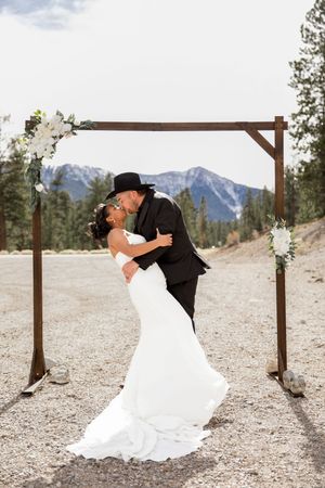 Romantic wedding couple dips and dances in front of rustic wooden arch with mountain backdrop in scenic outdoor setting.