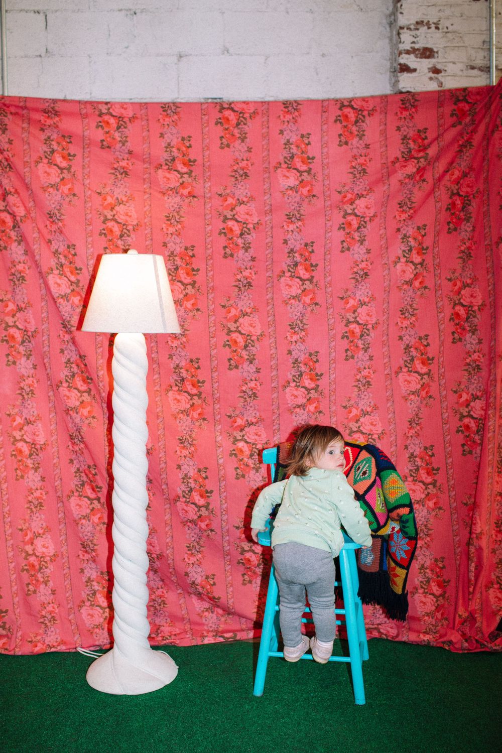 Child climbing turquoise stool against red floral backdrop with white floor lamp.