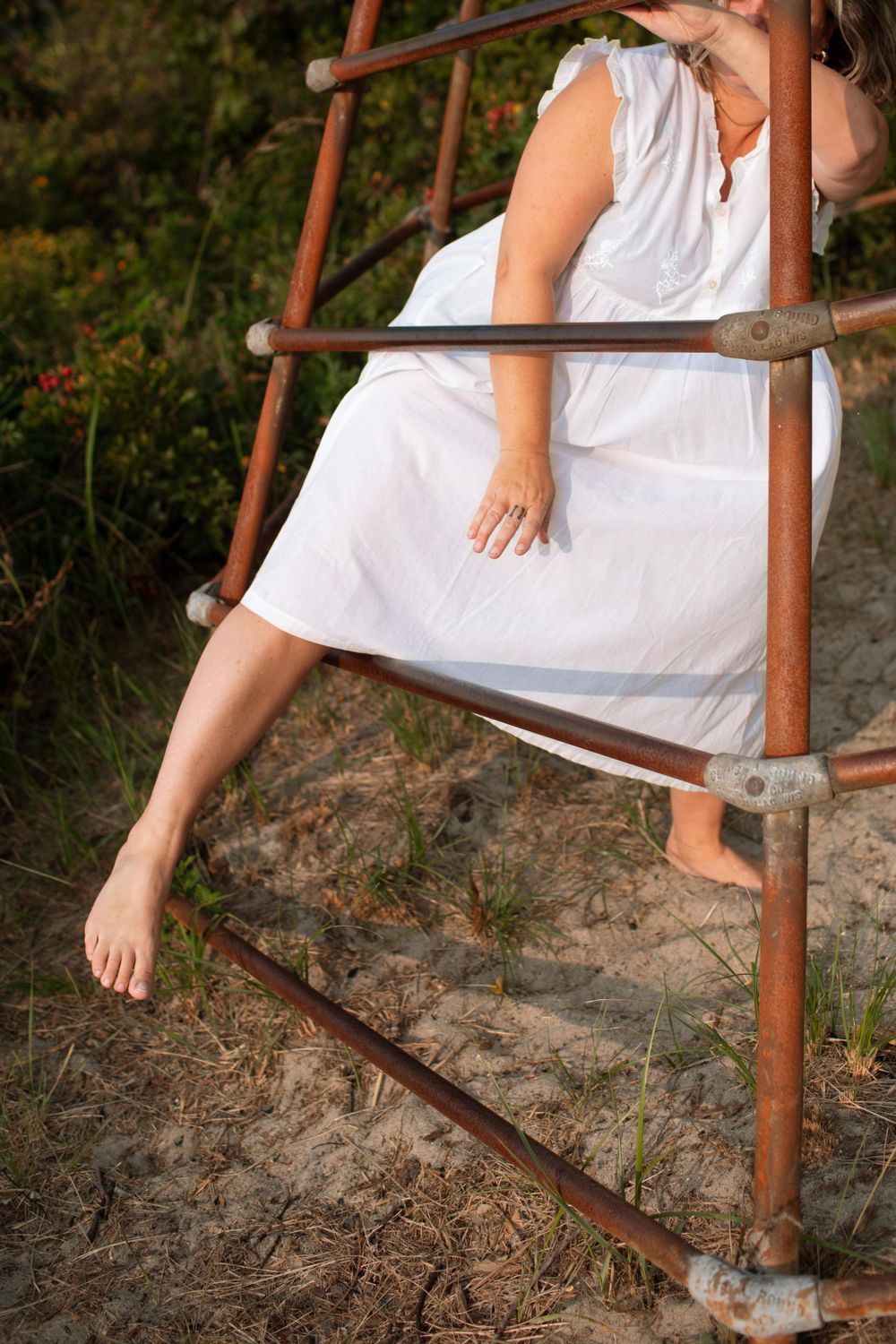 Close-up of bare feet and white dress on rustic metal ladder at beach.