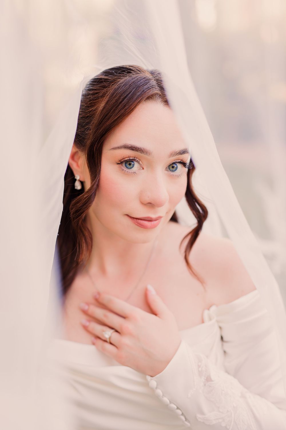 A bride with blue eyes peers through a white veil, wearing pearl earrings and an off-shoulder gown, hand on chest.