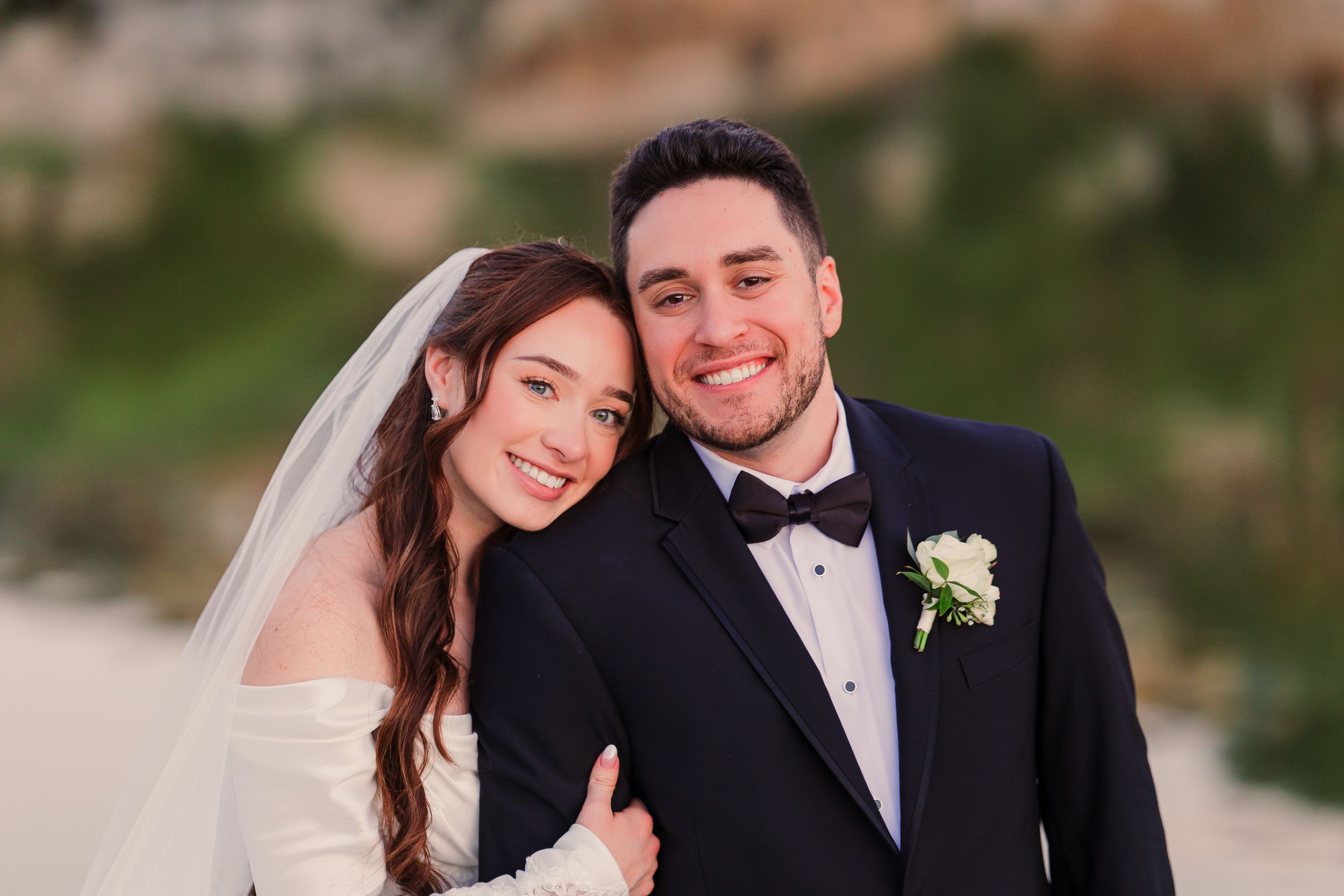 A smiling bride in a white gown and veil poses with a groom in a black tuxedo on a scenic outdoor wedding day.