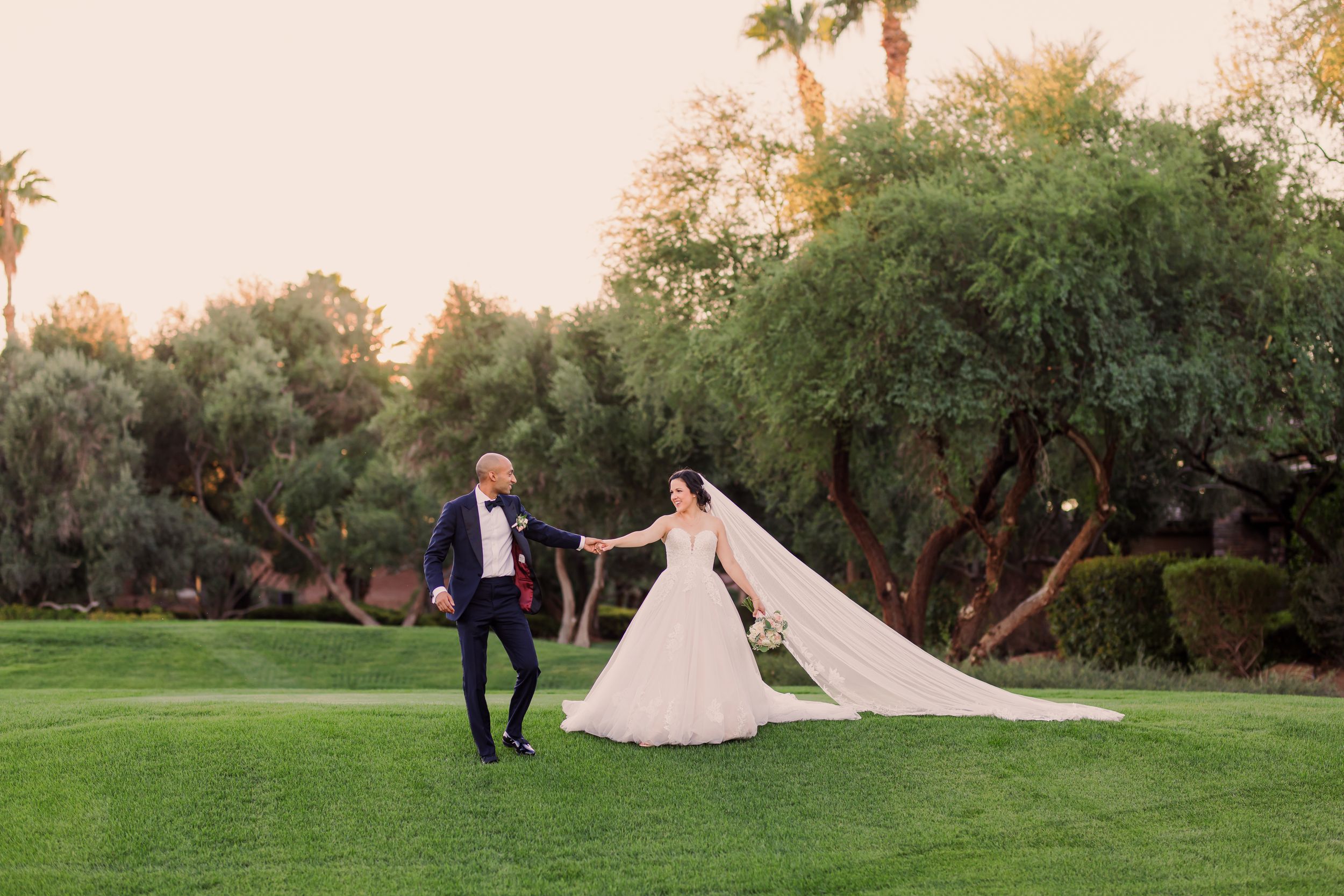 Bride and groom walk hand-in-hand on lush green golf course at sunset, bride's veil flowing behind her.