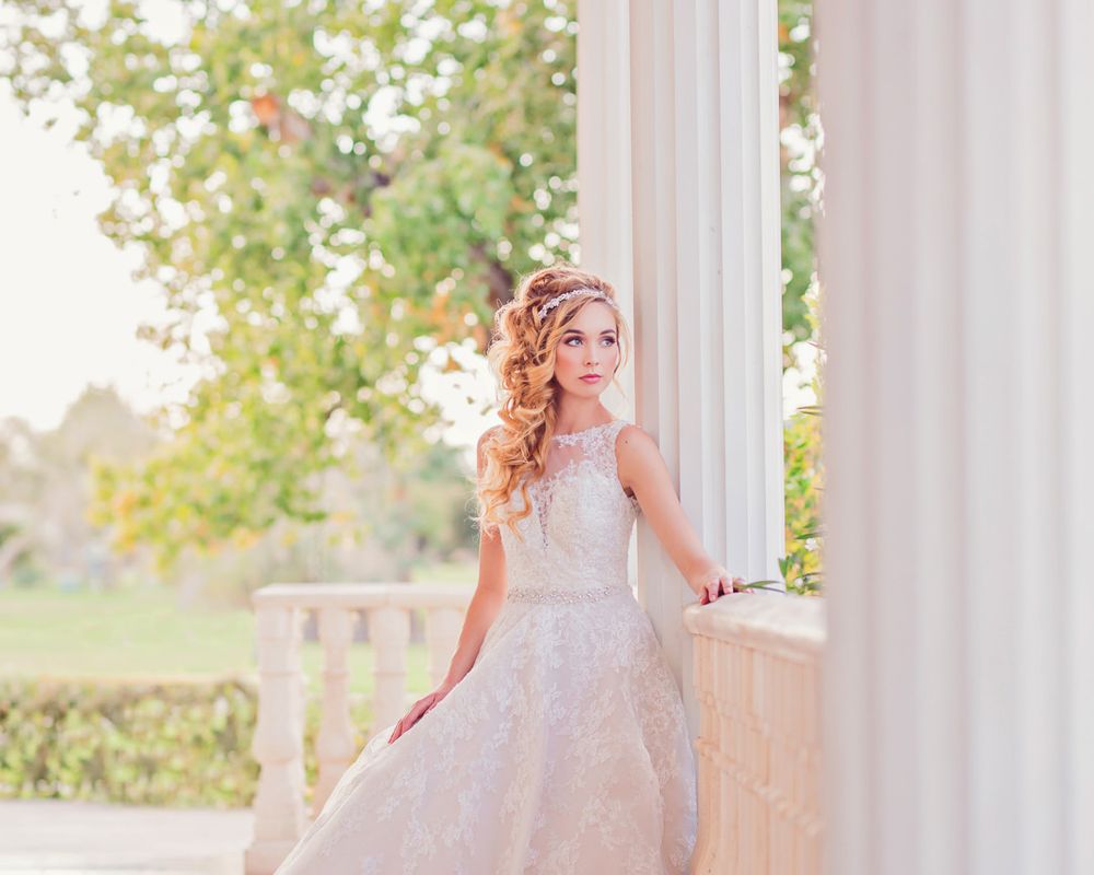 A bride in a flowing white wedding dress poses gracefully on an elegant outdoor porch with stone columns.