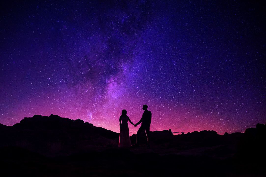 Silhouettes of two people stand beneath a vibrant purple night sky filled with stars and cosmic colors.