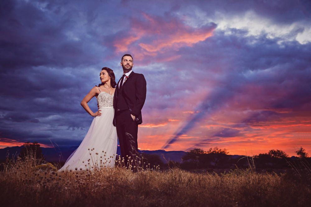 A couple stands dramatically against a vibrant sunset sky with dark storm clouds and mountain silhouettes.