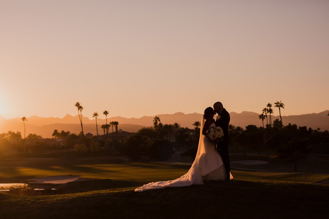 Silhouetted palm trees and figures against a warm orange sunset sky at a golf course.