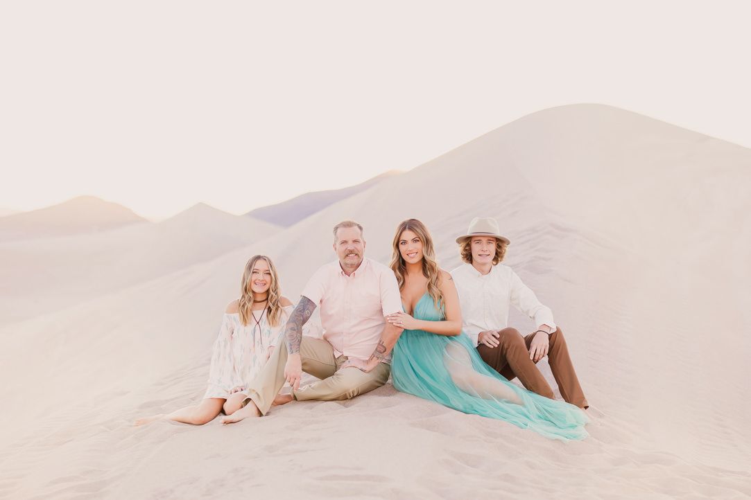A family sits together on sand dunes during sunset in light pastel-colored clothing.