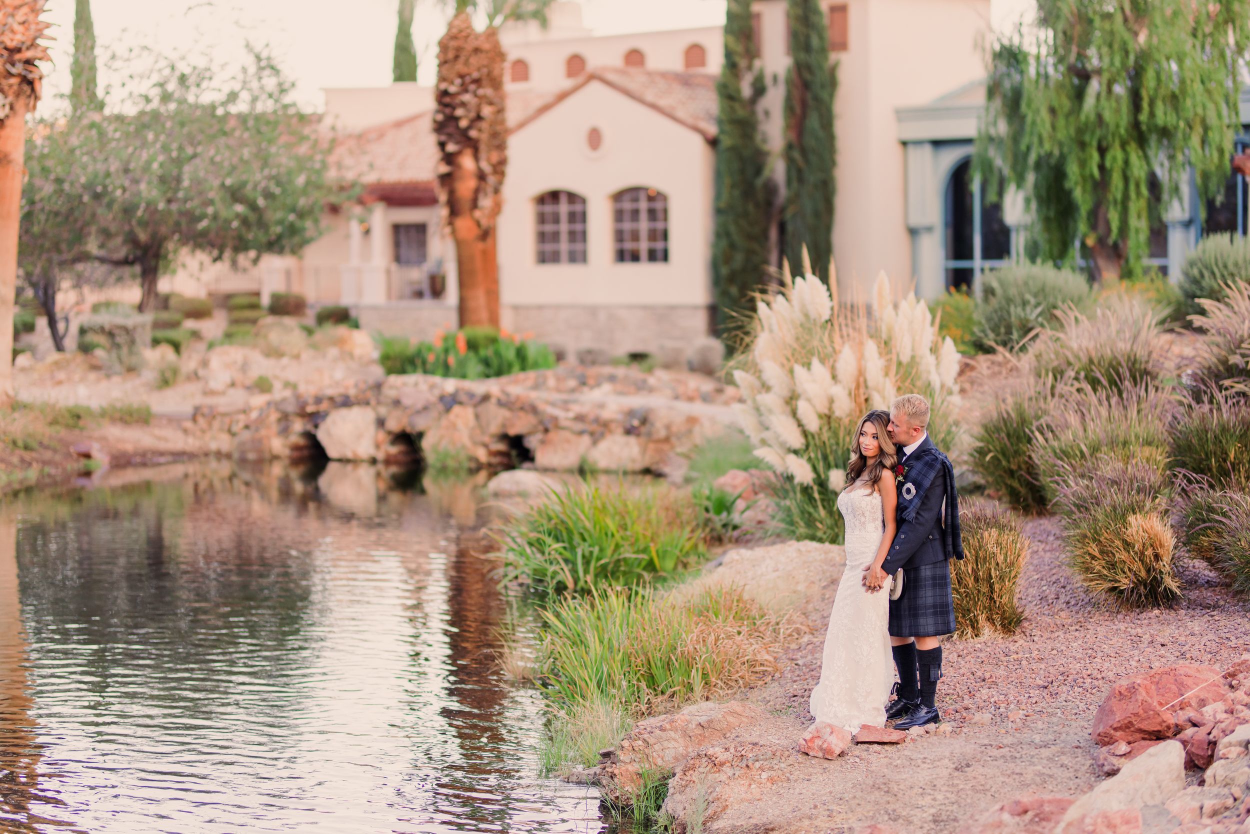 Bride and groom stand by a serene pond surrounded by desert landscaping and pampas grass at an elegant outdoor venue.