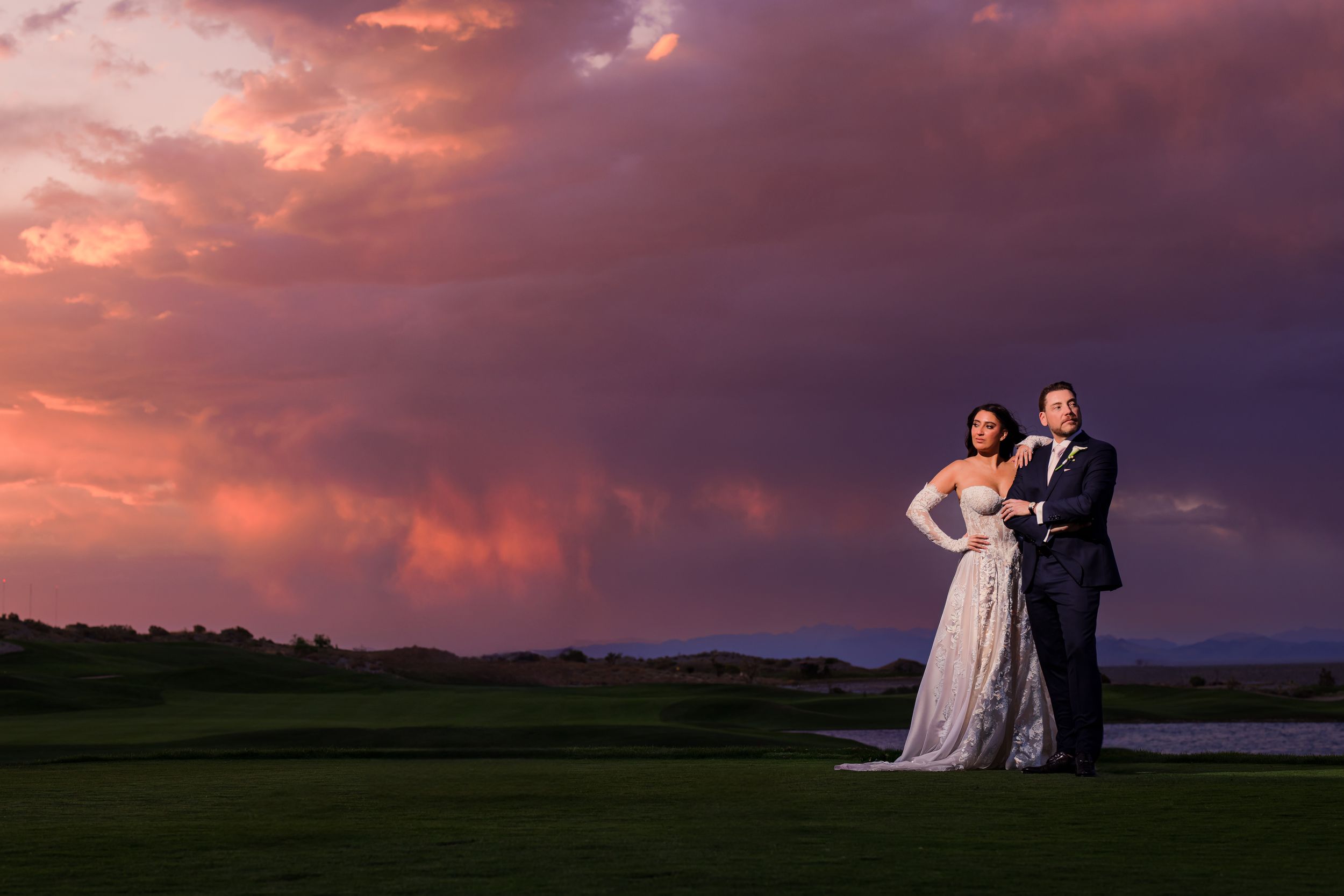 Bride and groom pose on a lush golf course under a dramatic pink and purple sunset sky.