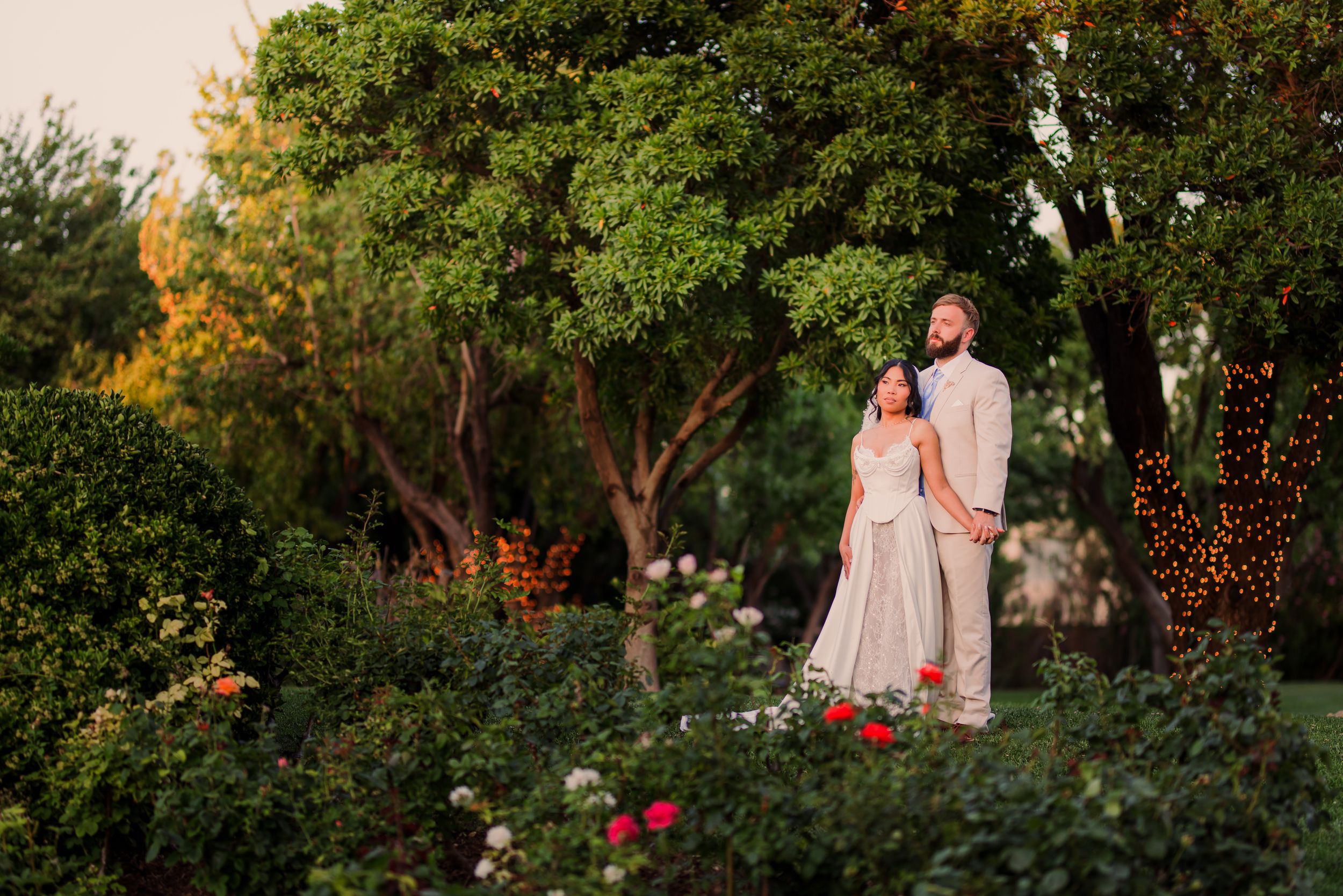 Couple in elegant attire embracing in a lush garden with roses and trees at golden hour.