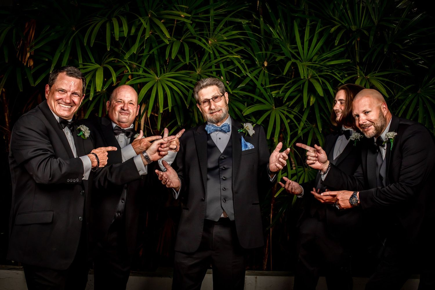 Wedding party members in formal attire stand laughing and toasting with champagne glasses against greenery backdrop.