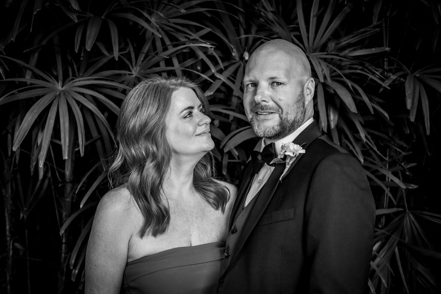 A black and white formal portrait of a couple in evening attire posing together against a palm leaf backdrop.