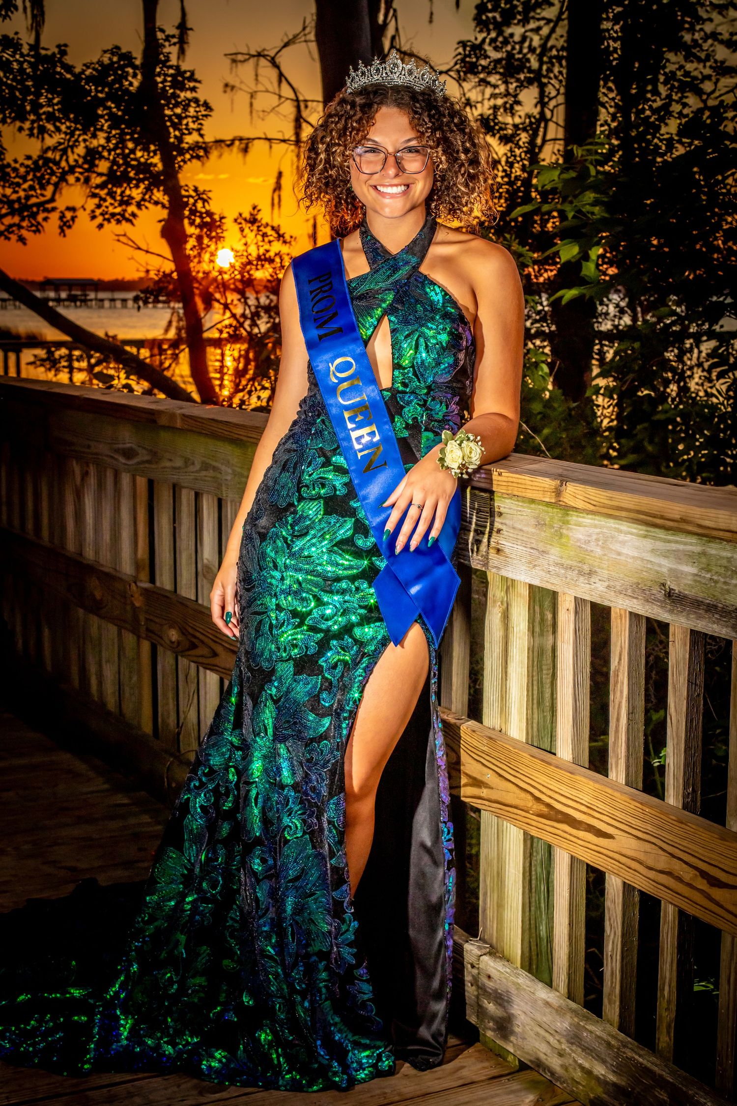 A person in a sparkly green formal gown and blue sash poses against a wooden railing at sunset creating a dramatic silhouette.