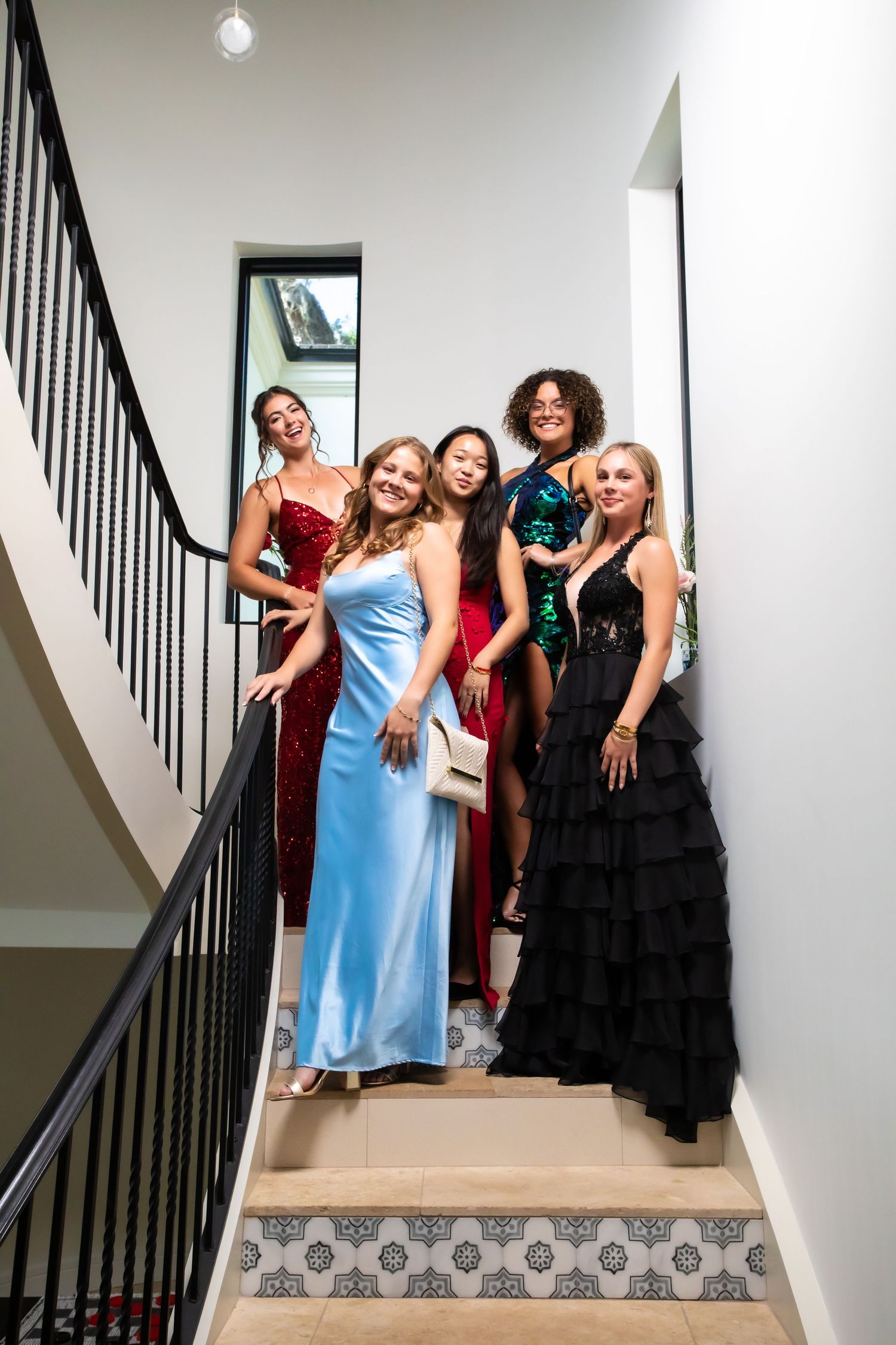 A group in formal attire poses together on an elegant staircase with white walls and black railings.