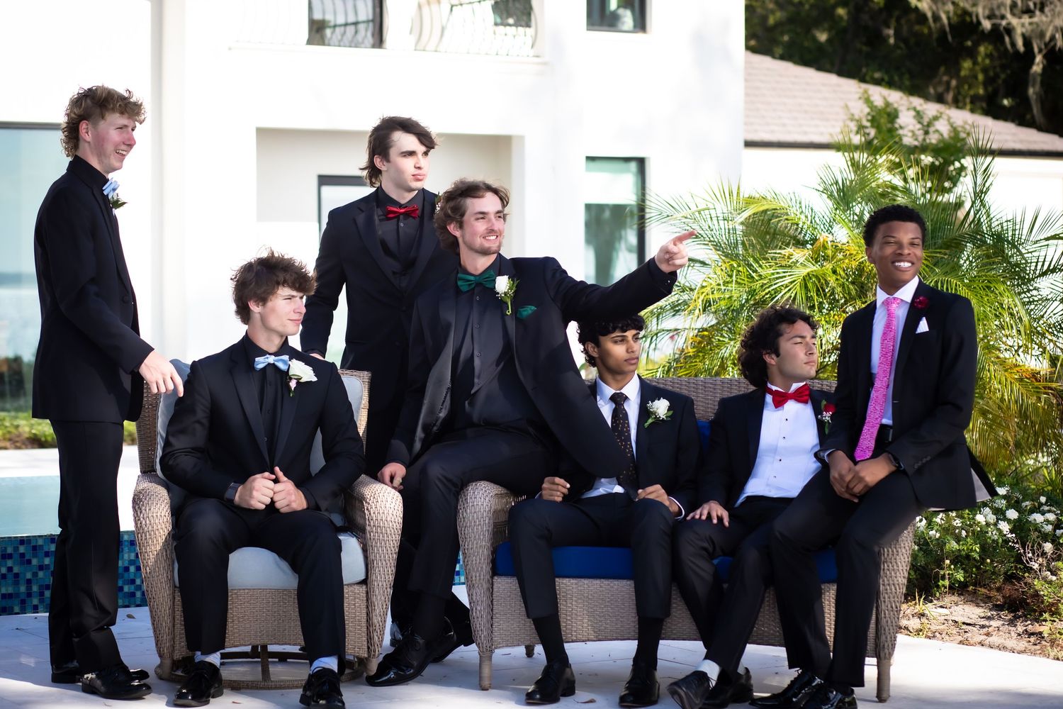 Group of groomsmen in black suits sit together on outdoor stairs near tropical plants.