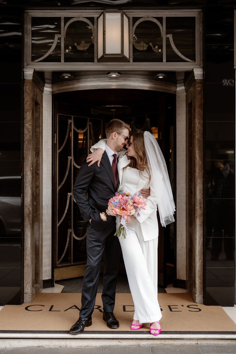 A couple in wedding attire share an intimate moment in an elegant doorway with pink floral bouquet and bright pink shoes.