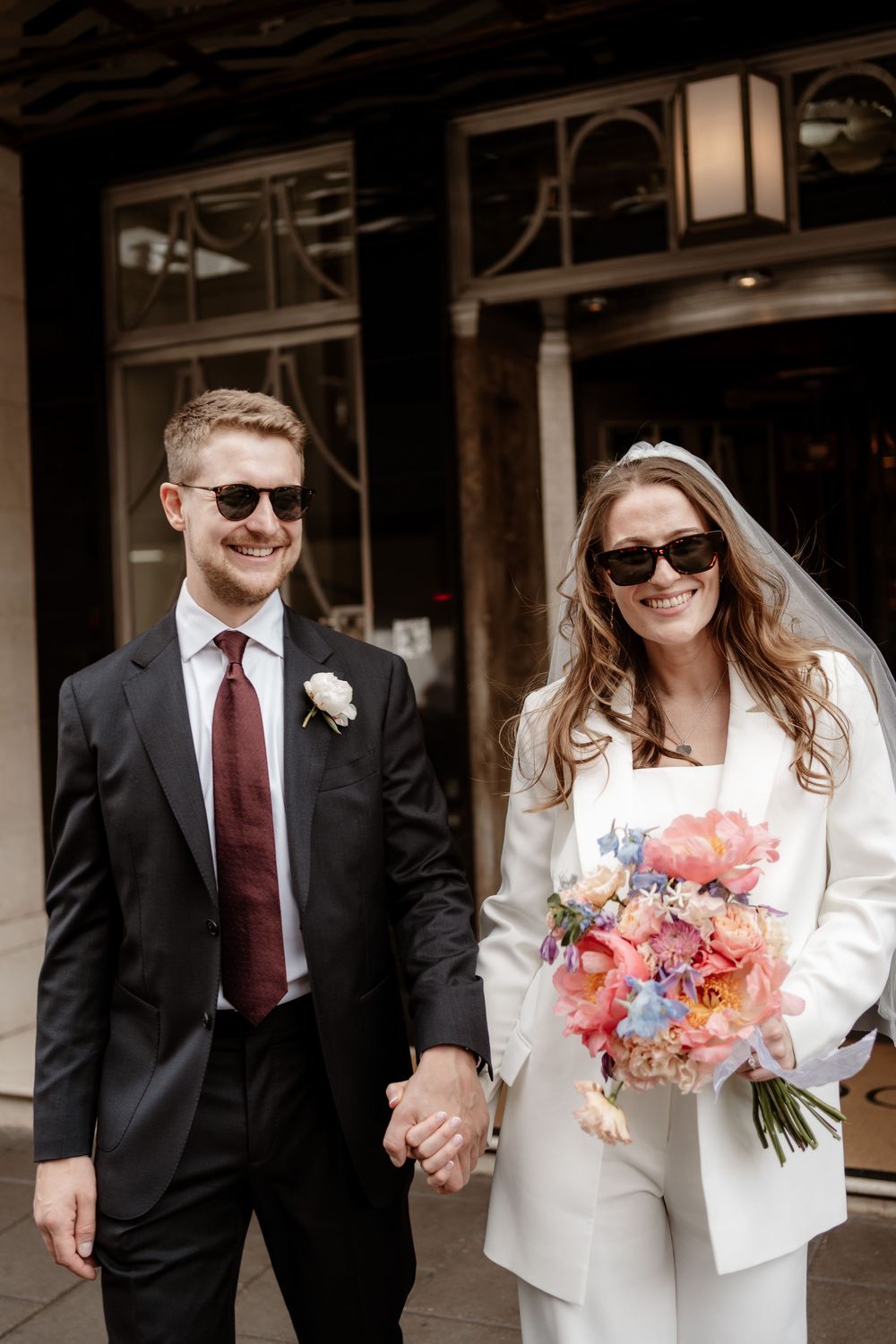 A stylish couple in sunglasses on their wedding day, with the bride holding a vibrant pink and coral bouquet.