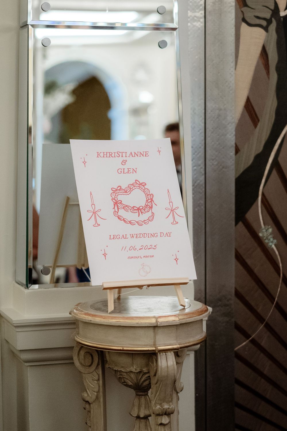 A pink wedding invitation with a wreath design sits propped on an ornate white pillar next to a mirror.
